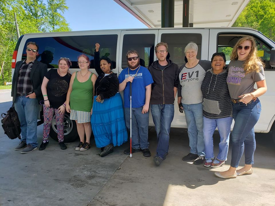 Down Home members gather outside a white van getting ready for a road trip. Quinterra Jones stands with fist raised above her head, wearing a blue skirt.