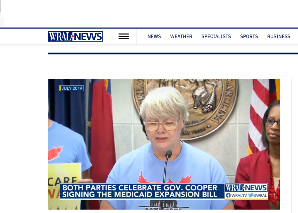 A woman wearing a Down Home NC t-shirt stands confidently at the podium, joining in the celebration of Governor Cooper signing the Medicaid Expansion bill, the image is captioned "both parties celebrate Governor Cooper signing the Medicaid Expansion bill"