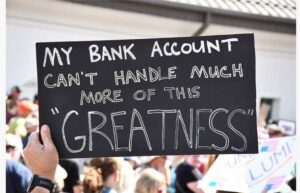 Photo of hands holding up a sign at a protest. In white writing on a black background, the sign says, "My bank account can't handle much more greatness."